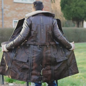 May include: A brown leather trench coat with a cream-colored collar and lining. The coat features multiple pockets, straps, and buckles. The back of the coat has stitched details and text. The person is standing outside on a sunny day.