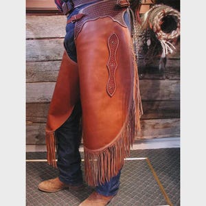 May include: Brown leather chaps with decorative stitching and fringe along the bottom edge. The chaps are worn over blue jeans and brown boots. A western wear item.