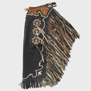 May include: Black leather chaps with tan and white floral embroidery and scrollwork. The chaps have long, layered fringe in brown, tan, and grey, and are accented with small leather details.