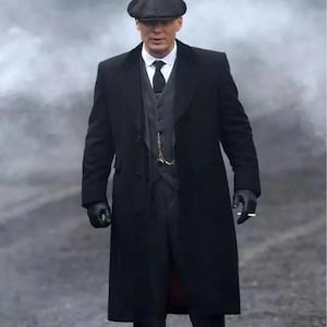 Puede incluir: Un hombre con un abrigo largo negro, chaleco y traje, que lleva una gorra plana oscura y guantes. Se ve una camisa blanca con cuello y una corbata negra. Una cadena de reloj de bolsillo cuelga del chaleco. El hombre sostiene un cigarrillo.
