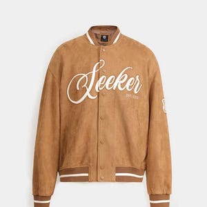 May include: A brown suede bomber jacket with the word "Seeker" in white script across the chest. The jacket has a ribbed collar, cuffs, and hem with white stripes. A small patch is on the upper right sleeve. The text "EST. 1999" is below the word "Seeker".