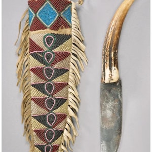 Puede incluir: Un cuchillo artesanal con mango de hueso y hoja de metal, junto a una funda de cuero con cuentas. La funda presenta un patrón geométrico en azul, rojo y negro, con flecos.
