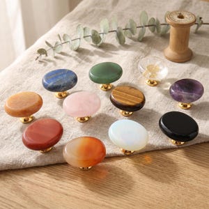 May include: A collection of eleven oval-shaped cabinet knobs featuring various polished gemstones and minerals, including blue, green, orange, pink, brown, purple, black, and clear stones, each mounted on a small, shiny gold-toned metal base.