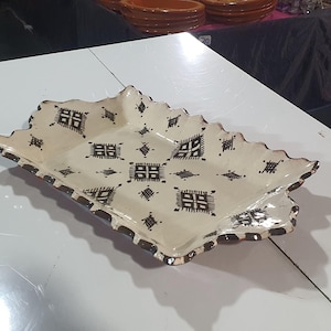 Könnte beinhalten: Ein rechteckiges, cremefarbenes Keramikserviertablett mit gewelltem Rand und braunem Rand. Das Tablett ist mit einem sich wiederholenden geometrischen Muster in Dunkelbraun verziert. Das Tablett steht auf einer weißen Oberfläche.