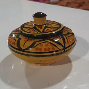 Könnte beinhalten: Ein kleiner, runder Keramiktopf mit Deckel. Der Topf ist mit einer gelben Basis, schwarzen und orangefarbenen geometrischen Mustern und einem dunkelbraunen Knopf auf dem Deckel bemalt. Der Topf steht auf einer weißen Oberfläche.