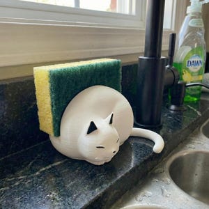 May include: A white cat-shaped sponge holder with black accents, holding a yellow and green sponge. The holder is on a dark countertop near a sink and faucet. A bottle of dish soap is visible in the background.