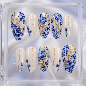 Op de afbeelding: Een set van tien ovale kunstnagels in een transparante doos. Elke nagel is beschilderd met een crèmewitte basis, versierd met blauwe bloemmotieven en gouden accenten. De nagels zijn in twee rijen van vijf gerangschikt.