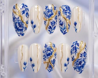May include: A set of ten oval-shaped press-on nails in a clear plastic case. Each nail is painted with a creamy white base, adorned with blue floral designs and gold accents. The nails are arranged in two rows of five.
