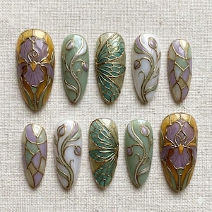 May include: Ten almond-shaped artificial nails with detailed designs. The nails display floral, butterfly wing, and stained glass patterns in gold, purple, green, and white. The designs are intricate and elegant, showcasing a variety of artistic styles.