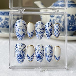 May include: A set of ten press-on nails featuring a blue and white porcelain design. Each nail has a white base with blue floral patterns, Pikachu illustrations, and gold accents. The nails are displayed in a clear plastic case.