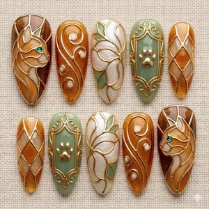 May include: A collection of ten almond-shaped nail art designs. The designs feature intricate gold outlines with various fillings, including a cat, floral patterns, a paw print, and geometric shapes. The color palette includes brown, gold, green, and white.