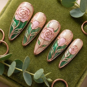 May include: Set of six almond-shaped nail art designs. Each nail features a pink rose design with green leaves, outlined in gold. The nails are displayed on a green velvet surface, with copper rings and greenery accents.