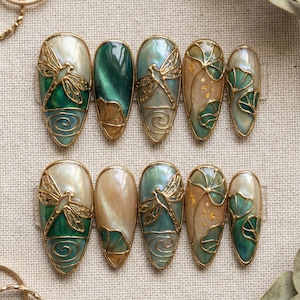 Puede incluir: Conjunto de diez uñas artificiales con diseño de libélula. Cada uña presenta un contorno dorado y una combinación de colores nacarados, verdes y dorados. La libélula es un elemento destacado, que añade un toque de elegancia inspirada en la naturaleza.