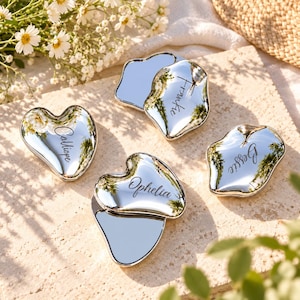 May include: Four silver-toned, reflective pocket mirrors with irregular, organic shapes. Each mirror is engraved with a name in a cursive script: Calliope, Frankie, Ophelia, and Bessie. The surfaces are highly polished and mirror-like.