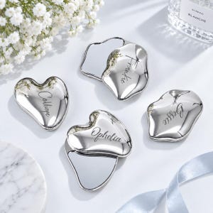 May include: Four silver-toned metal trinket dishes with irregular, organic shapes. Each dish is engraved with a name in a cursive script: Calliope, Frankie, Ophelia, and Bessie. The surfaces are highly reflective and polished to a mirror-like finish.