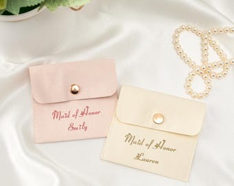 Könnte beinhalten: Zwei kleine, weiche Beutel in Hellrosa und Creme, jeweils mit einem goldenen Druckknopfverschluss. Der rosa Beutel ist mit "Maid of Honor Emily" beschriftet, der cremefarbene Beutel mit "Maid of Honor Lauren". Eine Perlenkette ist im Hintergrund.