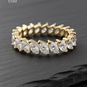 Könnte beinhalten: Ein goldener Ring mit einer durchgehenden Reihe von Diamanten im Marquiseschliff. Der Ring liegt auf einer dunklen Oberfläche. Der Ring ist von Bluepeak Exim.
