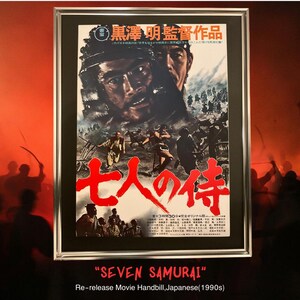 以下が含まれることがあります： 映画「七人の侍」のフレーム入りポスター。ポスターには、複数の男性のドラマチックなイメージが描かれており、赤と白の日本語のテキストが使用されています。ポスターは銀色のフレームに入っており、下部には「SEVEN SAMURAI」と「Re-release Movie Handbill, Japanese (1990s)」の文字があります。