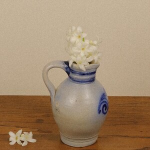 May include: A light grey ceramic pitcher with a single handle, featuring blue painted bands around the neck and a blue swirl design on the side. The pitcher contains a cluster of white flowers, and a small sprig of white flowers lies nearby.