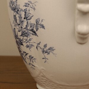 May include: A white ceramic jug featuring a blue floral pattern with detailed leaves and blossoms. The jug has a smooth, curved body and a textured base with a raised relief design. A vertical handle is visible on the right side.