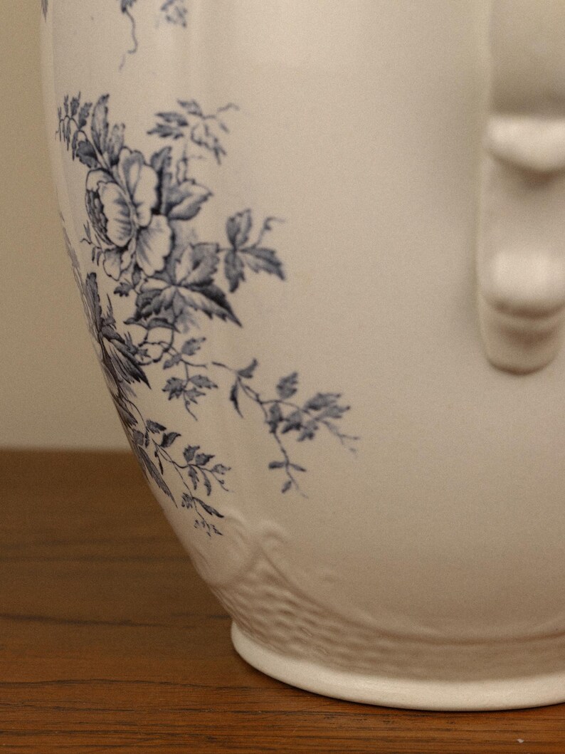 May include: A white ceramic jug featuring a blue floral pattern with detailed leaves and blossoms. The jug has a smooth, curved body and a textured base with a raised relief design. A vertical handle is visible on the right side.