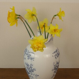 May include: A white ceramic jug featuring a blue floral pattern and a scalloped rim, containing a bouquet of bright yellow daffodils with green stems. The jug has a rounded body and a handle, displaying a classic transferware design.