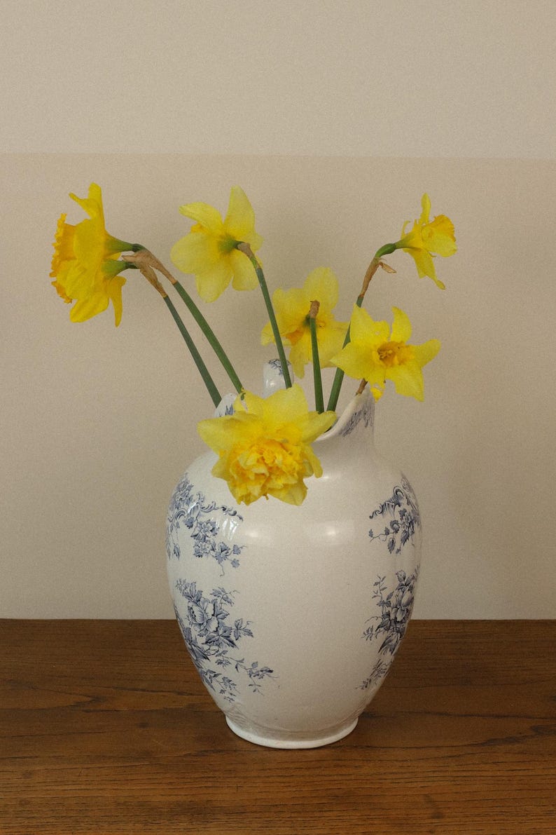 May include: A white ceramic jug featuring a blue floral pattern and a scalloped rim, containing a bouquet of bright yellow daffodils with green stems. The jug has a rounded body and a handle, displaying a classic transferware design.