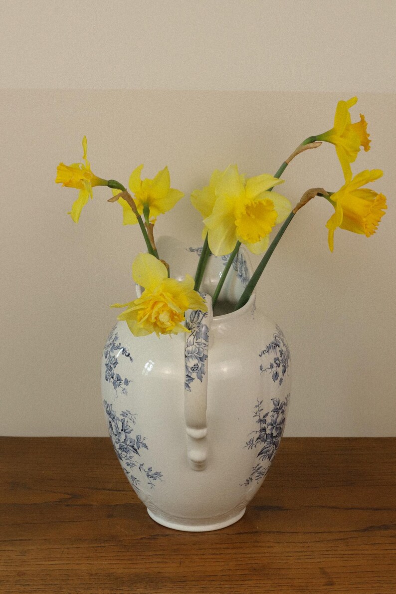May include: A white ceramic jug featuring a blue floral pattern and a curved handle, containing a bouquet of bright yellow daffodils with green stems. The jug has a rounded body and a flared rim, displaying detailed blue botanical illustrations.