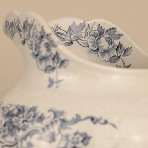 May include: A white ceramic jug featuring a scalloped rim and a detailed blue floral pattern. The design includes intricate illustrations of roses and leaves wrapping around the upper body and the curved spout of the vessel.
