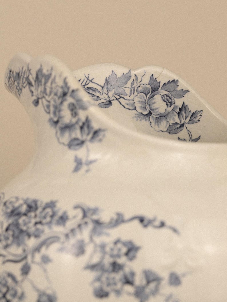 May include: A white ceramic jug featuring a scalloped rim and a detailed blue floral pattern. The design includes intricate illustrations of roses and leaves wrapping around the upper body and the curved spout of the vessel.
