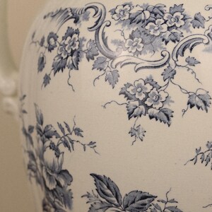May include: A white ceramic jug featuring a blue floral pattern with intricate botanical illustrations of flowers, leaves, and scrolling vines. The surface displays a detailed transferware design covering the curved body of the vessel.