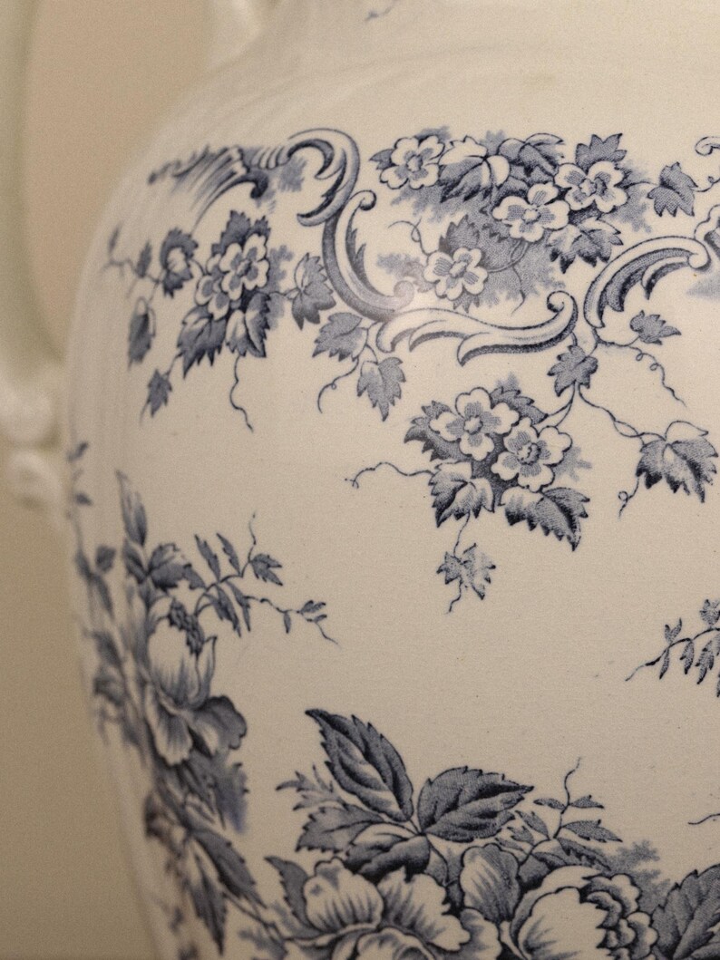May include: A white ceramic jug featuring a blue floral pattern with intricate botanical illustrations of flowers, leaves, and scrolling vines. The surface displays a detailed transferware design covering the curved body of the vessel.