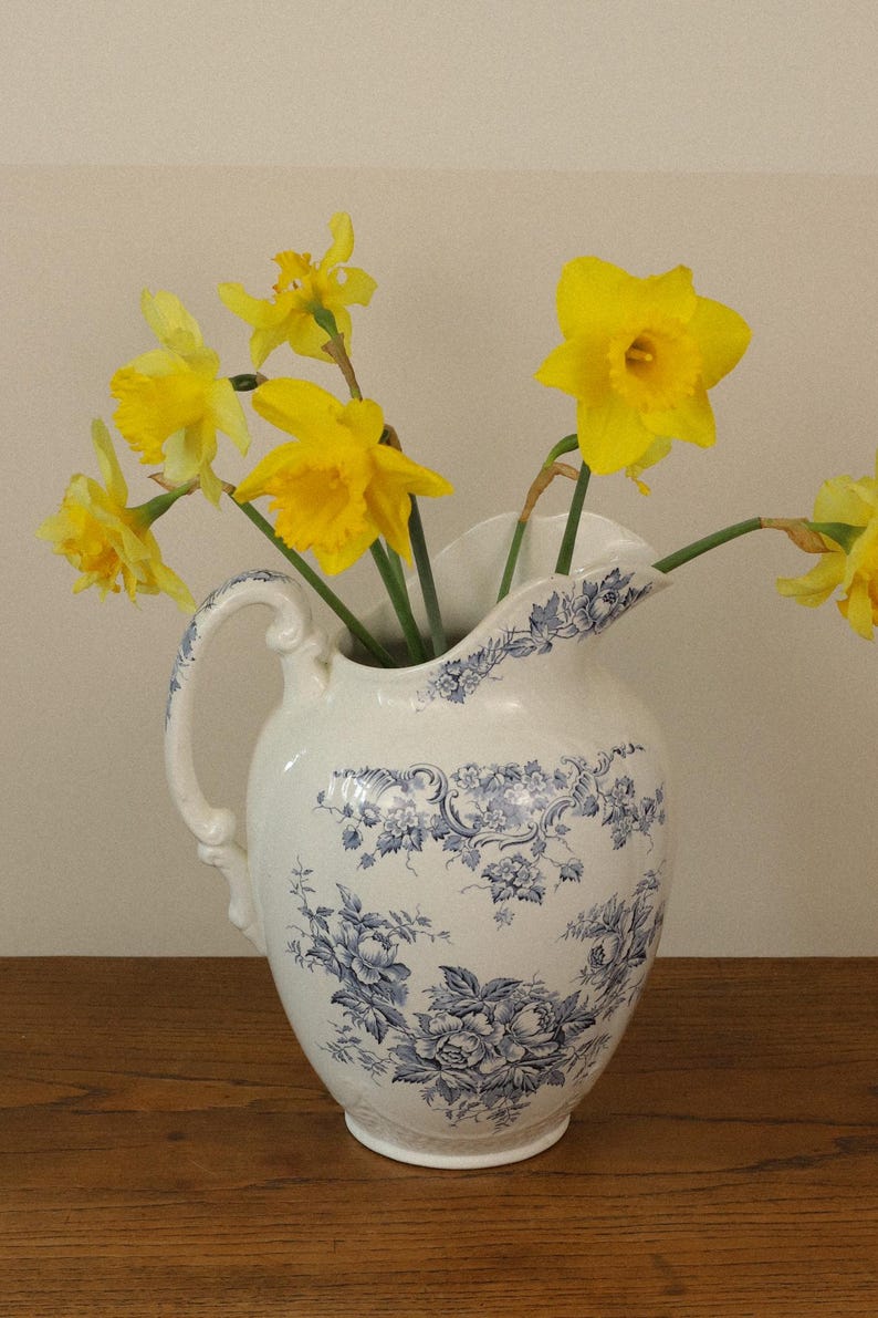 May include: A white ceramic jug featuring a blue floral pattern and a curved handle, filled with a bouquet of bright yellow daffodils. The jug has a wide base and a decorative spout, showcasing intricate botanical illustrations in blue ink.