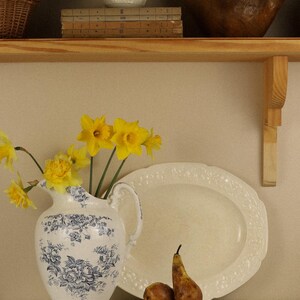 May include: A white ceramic jug with blue floral patterns holds bright yellow daffodils. Beside it sits a large oval cream-coloured platter, two brown pears on a wooden cutting board, and a white lace tablecloth with floral embroidery.