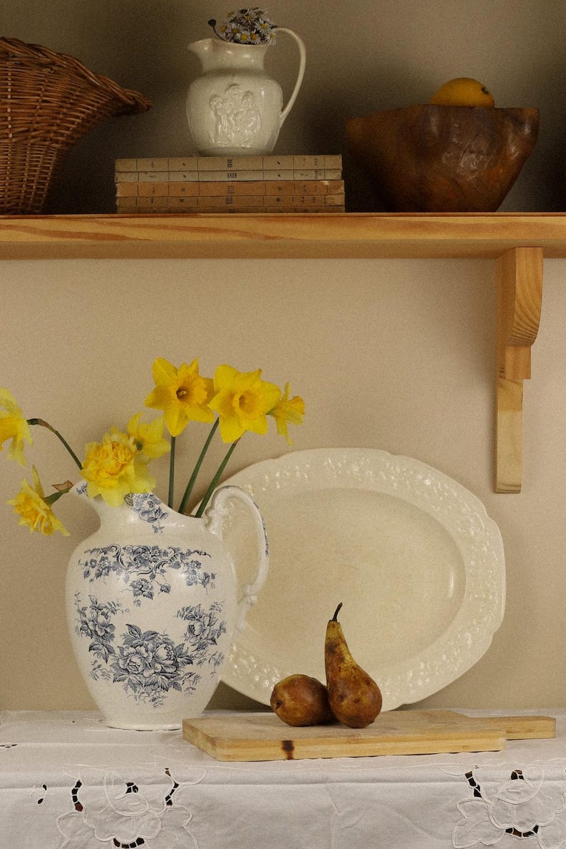 May include: A white ceramic jug with blue floral patterns holds bright yellow daffodils. Beside it sits a large oval cream-coloured platter, two brown pears on a wooden cutting board, and a white lace tablecloth with floral embroidery.