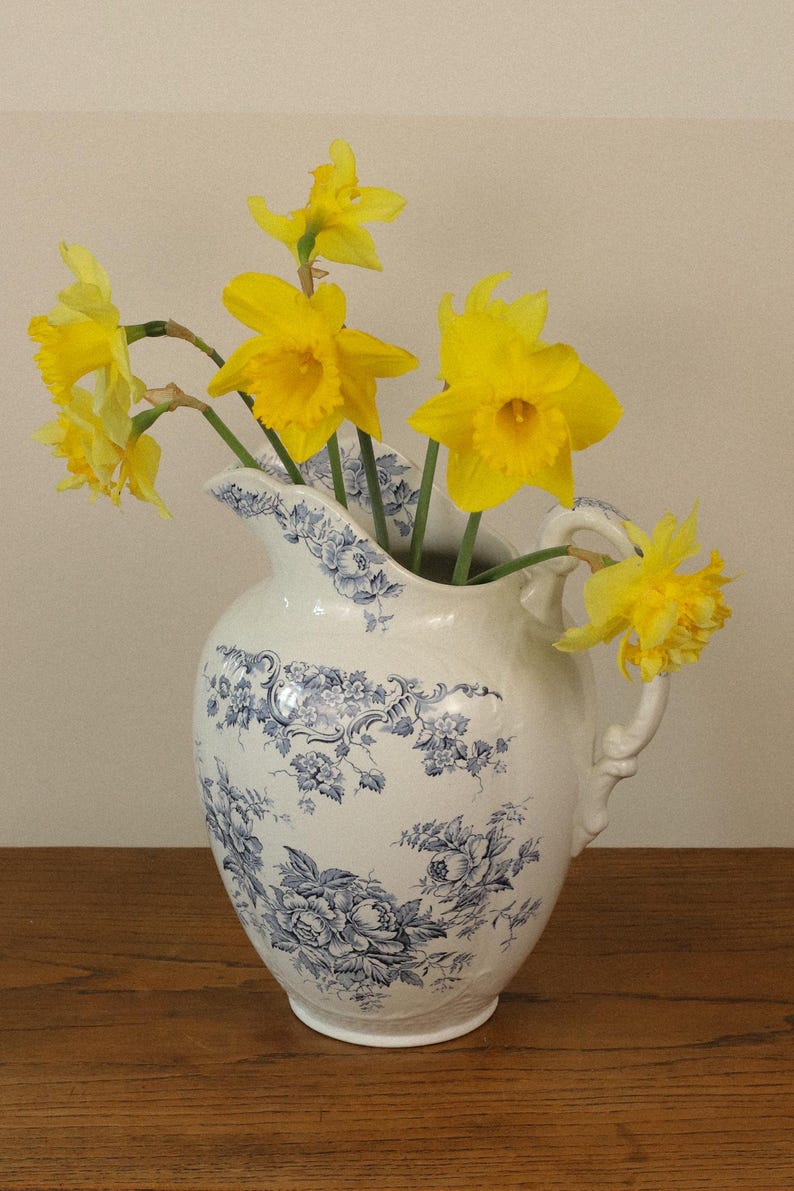 May include: A white ceramic jug featuring a blue floral pattern with a curved handle and a scalloped rim. The jug is filled with several bright yellow daffodils with green stems, with one flower resting near the handle.