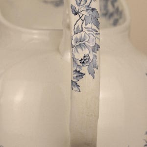 May include: A white ceramic jug featuring a curved handle decorated with a blue floral pattern. The design includes detailed blue leaves and blossoms running along the length of the handle, set against a smooth, off-white glazed surface.