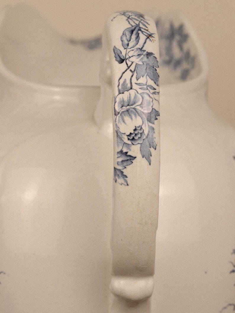 May include: A white ceramic jug featuring a curved handle decorated with a blue floral pattern. The design includes detailed blue leaves and blossoms running along the length of the handle, set against a smooth, off-white glazed surface.