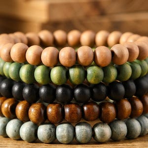 May include: Close-up of several stacked wooden bead bracelets in various colours. The bracelets include natural wood, green, black, brown, and grey beads. The beads are round and appear to be strung on elastic or cord.