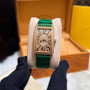 Op de afbeelding: Een rechthoekig gouden polshorloge met een groene leren band. De wijzerplaat van het horloge is bezet met kleine steentjes en heeft zwarte cijfers. Het horloge wordt gepresenteerd op een cr&egrave;mekleurig oppervlak.