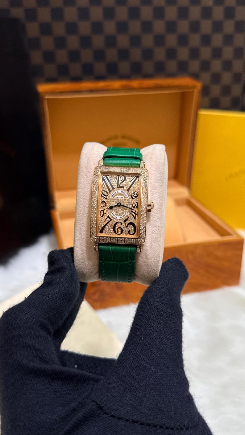 Op de afbeelding: Een rechthoekig gouden polshorloge met een groene leren band. De wijzerplaat van het horloge is bezet met kleine steentjes en heeft zwarte cijfers. Het horloge wordt gepresenteerd op een cr&egrave;mekleurig oppervlak.