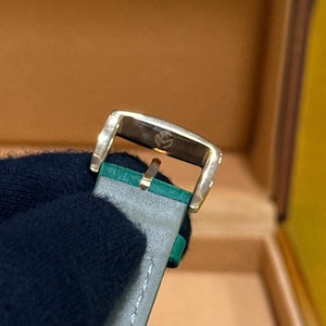 Op de afbeelding: Close-up van een goudkleurige horlogegesp met een grijze leren band en groene accenten. De gesp heeft een klein embleem. De horlogeband is gestikt met witte draad. De achtergrond is een bruine doos.