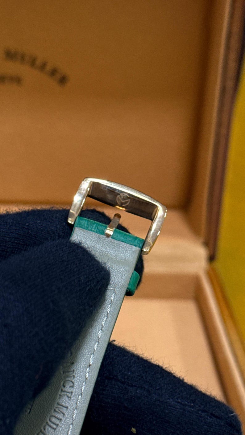 Op de afbeelding: Close-up van een goudkleurige horlogegesp met een grijze leren band en groene accenten. De gesp heeft een klein embleem. De horlogeband is gestikt met witte draad. De achtergrond is een bruine doos.