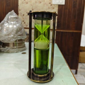 Puede incluir: Un reloj de arena de estilo vintage con marco de latón y vidrio verde. La arena es de color beige claro. El reloj de arena es un objeto decorativo, posiblemente para cronometrar o exhibir. El diseño es clásico y atemporal. Altura aproximada 15 cm.