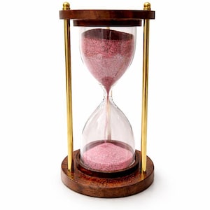 May include: Hourglass with a wooden frame and base, featuring clear glass chambers and pink sand. The hourglass has gold-colored metal supports and is set against a white background. A classic decorative item.