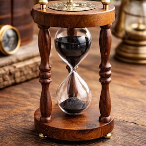 Op de afbeelding: Een houten zandloper met een heldere glazen bol gevuld met zwart zand. De zandloper heeft een donkerbruin houten frame met messing accenten. De zandloper is een decoratief item.