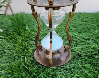 Può includere: Una clessidra antica in ottone con una camera di vetro trasparente riempita di sabbia blu. La clessidra ha una parte superiore e una base circolari decorative, sostenute da gambe curve ornate. La parte superiore è incisa con testo. La clessidra è su erba verde.