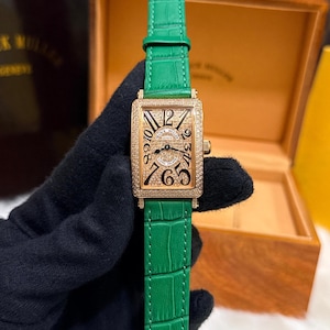 Op de afbeelding: Een gouden rechthoekig polshorloge met een groene leren band. De wijzerplaat van het horloge heeft zwarte cijfers en een met diamanten bezette lunette. Het horloge wordt vastgehouden door een gehandschoende hand, met een houten doos op de achtergrond.