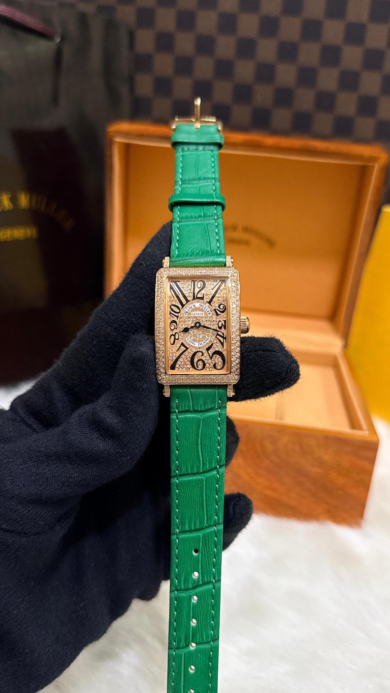 Op de afbeelding: Een gouden rechthoekig polshorloge met een groene leren band. De wijzerplaat van het horloge heeft zwarte cijfers en een met diamanten bezette lunette. Het horloge wordt vastgehouden door een gehandschoende hand, met een houten doos op de achtergrond.