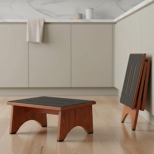 May include: A wooden step stool with a black top and brown legs. The stool is shown in two positions: assembled and folded. The assembled stool is on the floor, while the folded stool leans against a wall. The background is a kitchen.